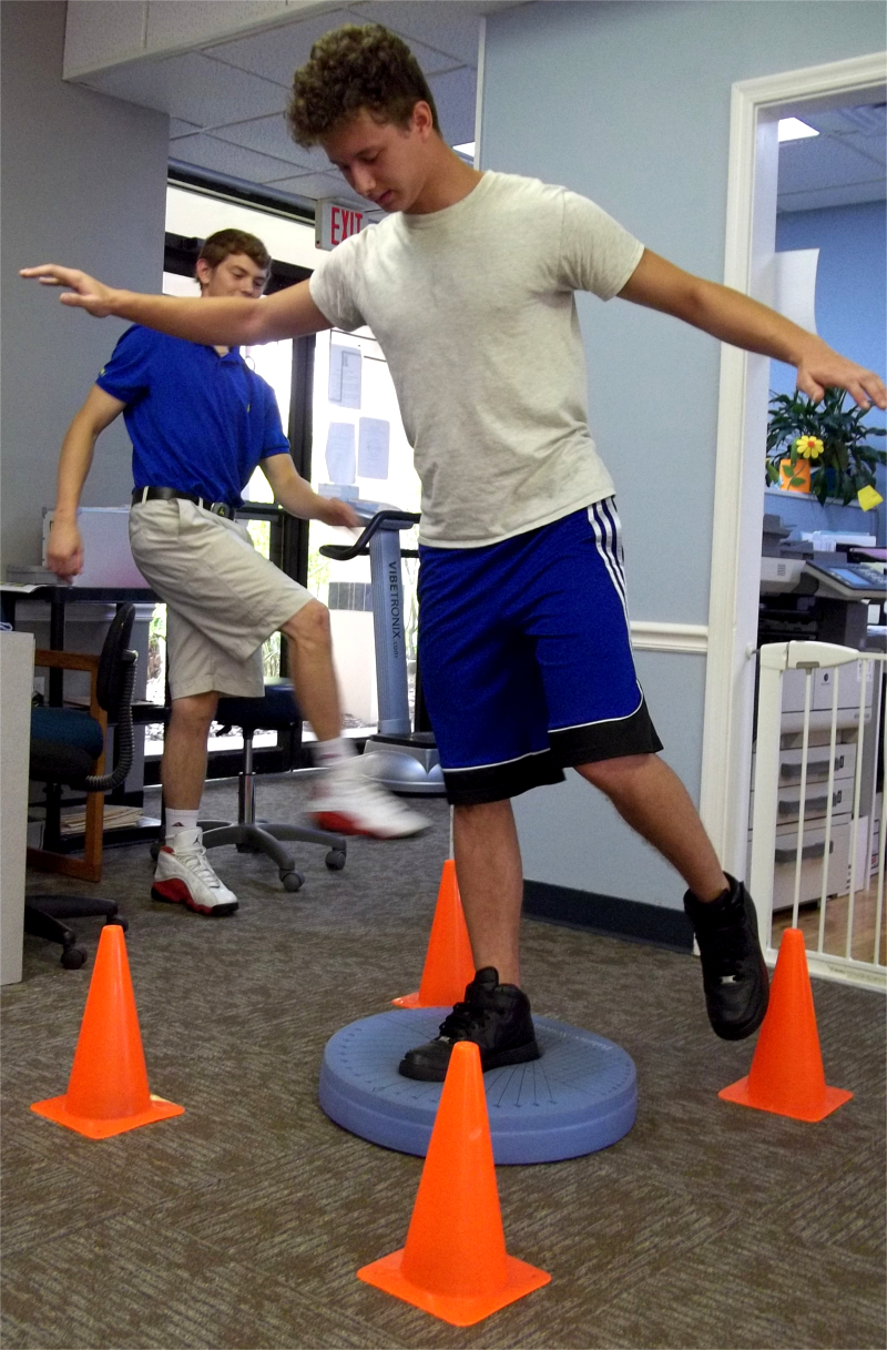 Young Man, Balancing one Foot on Soft Surface and Touch Toe to Orange Cone Behind Him. Three other Cones Place Around Him as well.